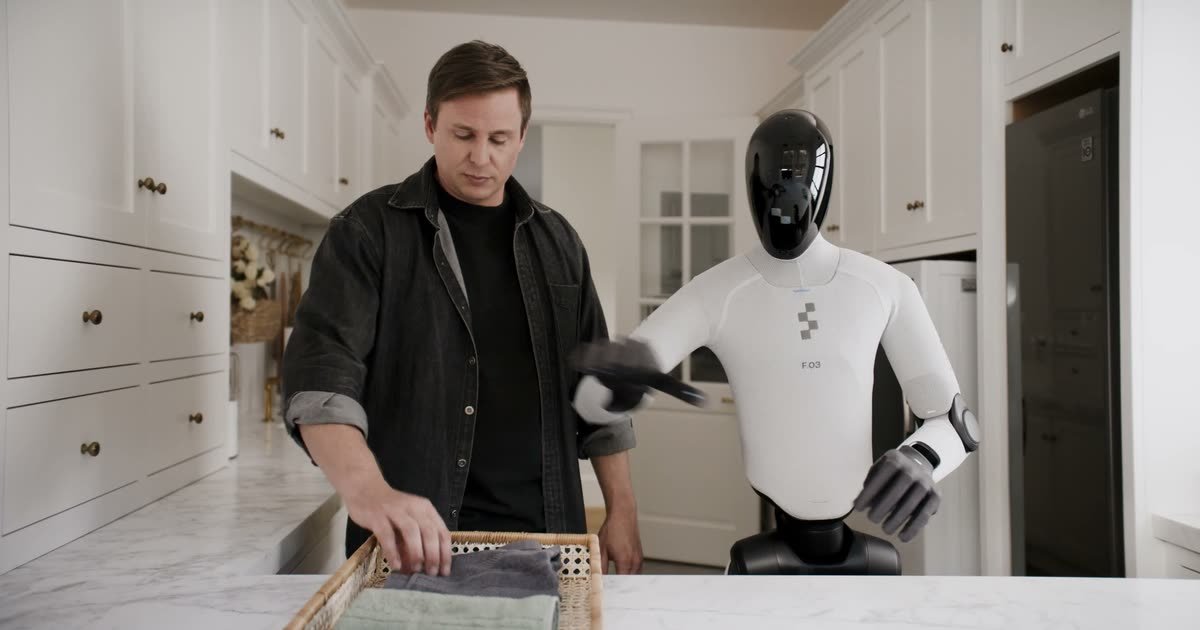 Robot Figure 03 z napisem F.03 na piersi składa ręcznik przy blacie, obok Brett Adcock sięga do koszyka z ręcznikami